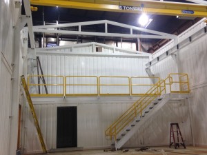 Mezzanine with yellow guard rail and steel staircase, also with yellow guard rail in between white steel walls.