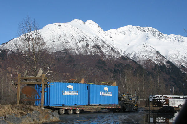 Two sky blue fabricated shipping containers loaded on flat trailer. Containers have white STEELFAB logo and white outline of a bison
